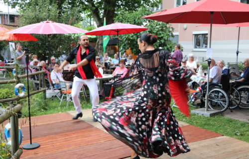 Ein Paar tanzt Flamenco auf einer Bühne im Freien, umgeben von Zuschauern unter roten Sonnenschirmen.