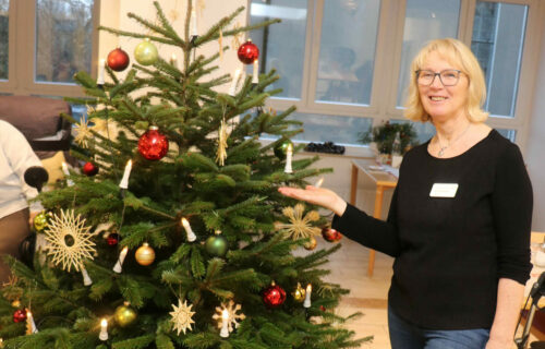 Eine Frau steht neben einem geschmückten Weihnachtsbaum in einem Raum mit großen Fenstern im Hintergrund.