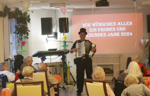 Ein Mann mit Akkordeon spielt vor Senioren. Im Hintergrund ein Bildschirm mit Neujahrsgrüßen für 2024. Luftballons dekorieren den Raum.
