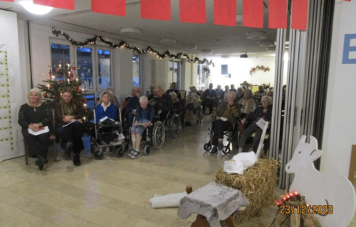 Ältere Menschen sitzen in Rollstühlen in einem festlich dekorierten Raum mit Weihnachtsbaum und Krippenfiguren.