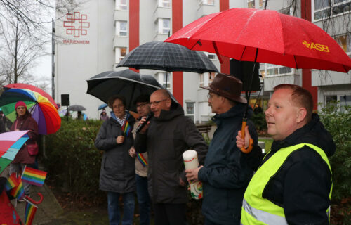 Personen unter Regenschirmen bei einer Veranstaltung vor einem Gebäude, eine Person hält ein Mikrofon.