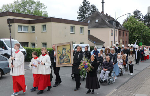 Prozession mit Ikone und Blumen in einer Straße; Menschen in liturgischer Kleidung und Begleitung von Rollstühlen.