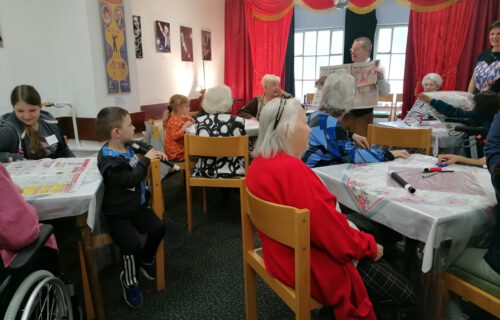 Gruppe von Senioren und Kindern in einem Gemeinschaftsraum, umgeben von roten Vorhängen, während eines interaktiven Treffens.