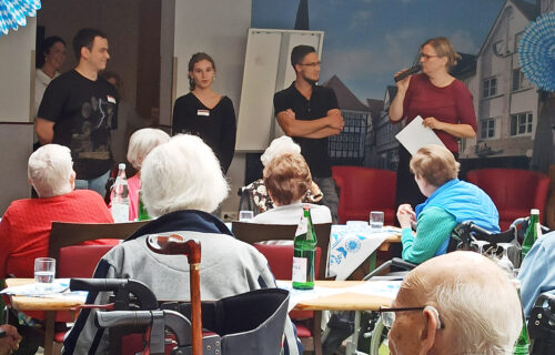 Veranstaltung in einem Seniorenheim: Eine Frau spricht zu drei stehenden Personen, Senioren sitzen an Tischen im Vordergrund.