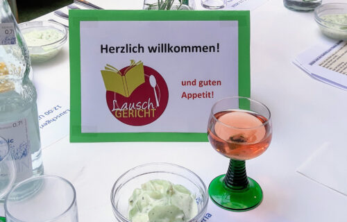Willkommensschild bei Lauschgericht-Event mit Getränk, grüner Sauce und Menükarten auf einem Tisch.