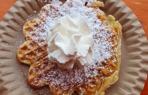 Frische Waffel mit Puderzucker und Sahne auf Pappteller, serviert auf einem Holz-Tisch. Perfekt für den süßen Genuss!