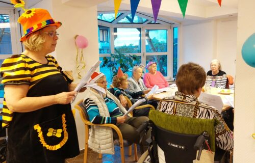 Ältere Menschen feiern in bunten Kostümen eine Party mit Singen und Dekoration in einem Gemeinschaftsraum.